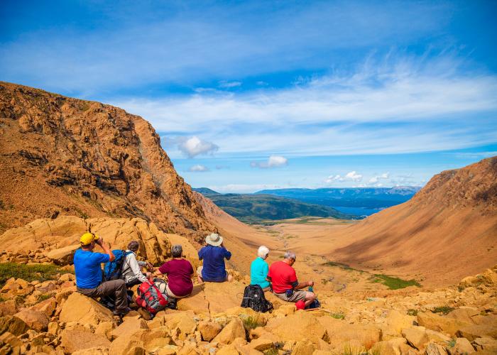 A group of six older hikers take a moment to rest and take in the scenery on the tablelands at Gros Morne National Park. 