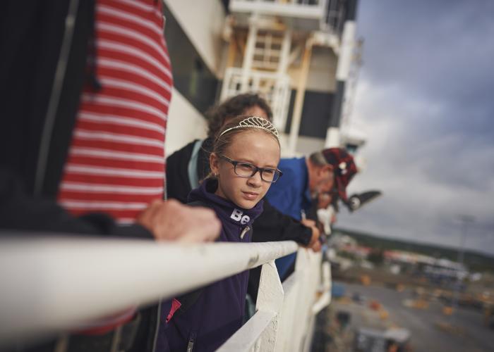 A young girl wearing a tiara and her family look over the white rails of a Marine Atlantic ferry as it leaves port. 