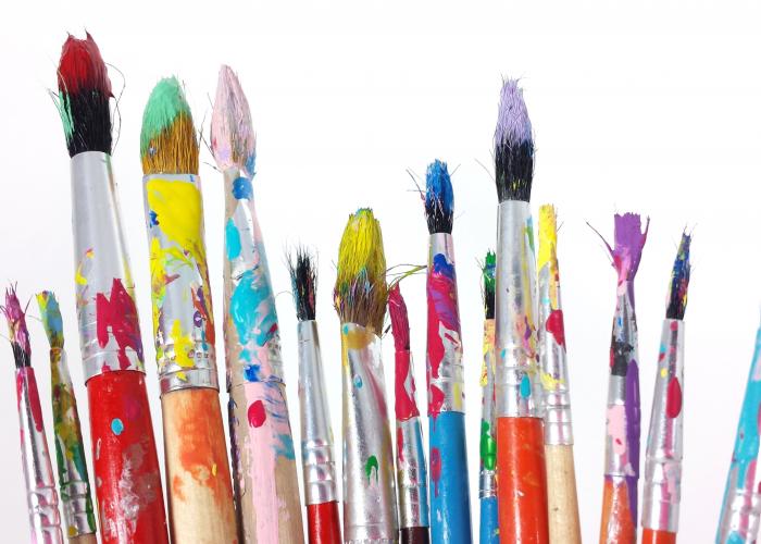 A grouping of art paintbrushes in various sizes, covered in splattered paint colours and set against a white background.
