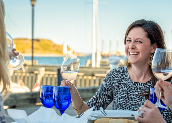 A smiling brunette woman raises a glass of white wine during a patio dinner at a restaurant on the Halifax waterfront.