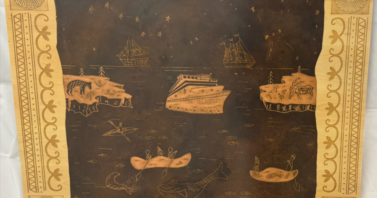 Engraved wooden plaque on the Ala'suinu vessel, featuring Mi'kmaw-style artwork of ships, canoes, wildlife, and stars, with an inscription explaining the vessel's name.