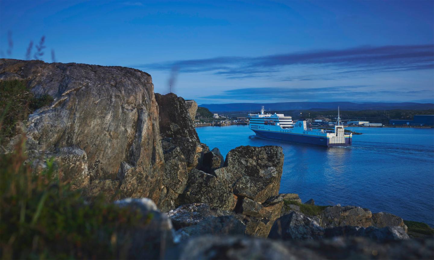A large white and blue Marine Atlantic ferry arriving at a rocky harbour port at dusk.