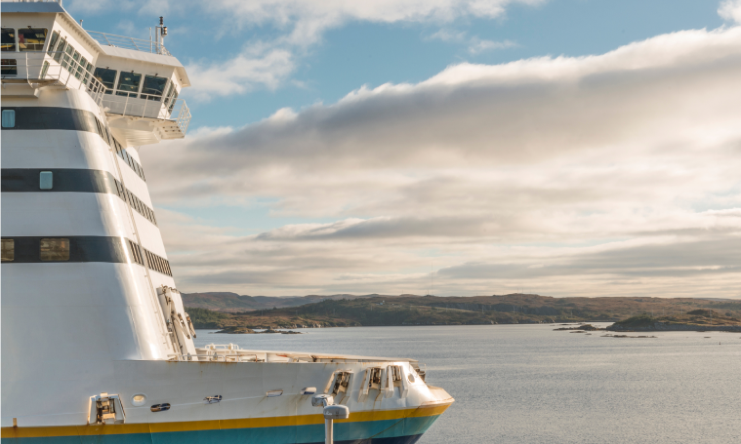 The bow and upper decks of a ferry vessel docked at a calm harbour, with rolling hills and a partly cloudy sky in the background.