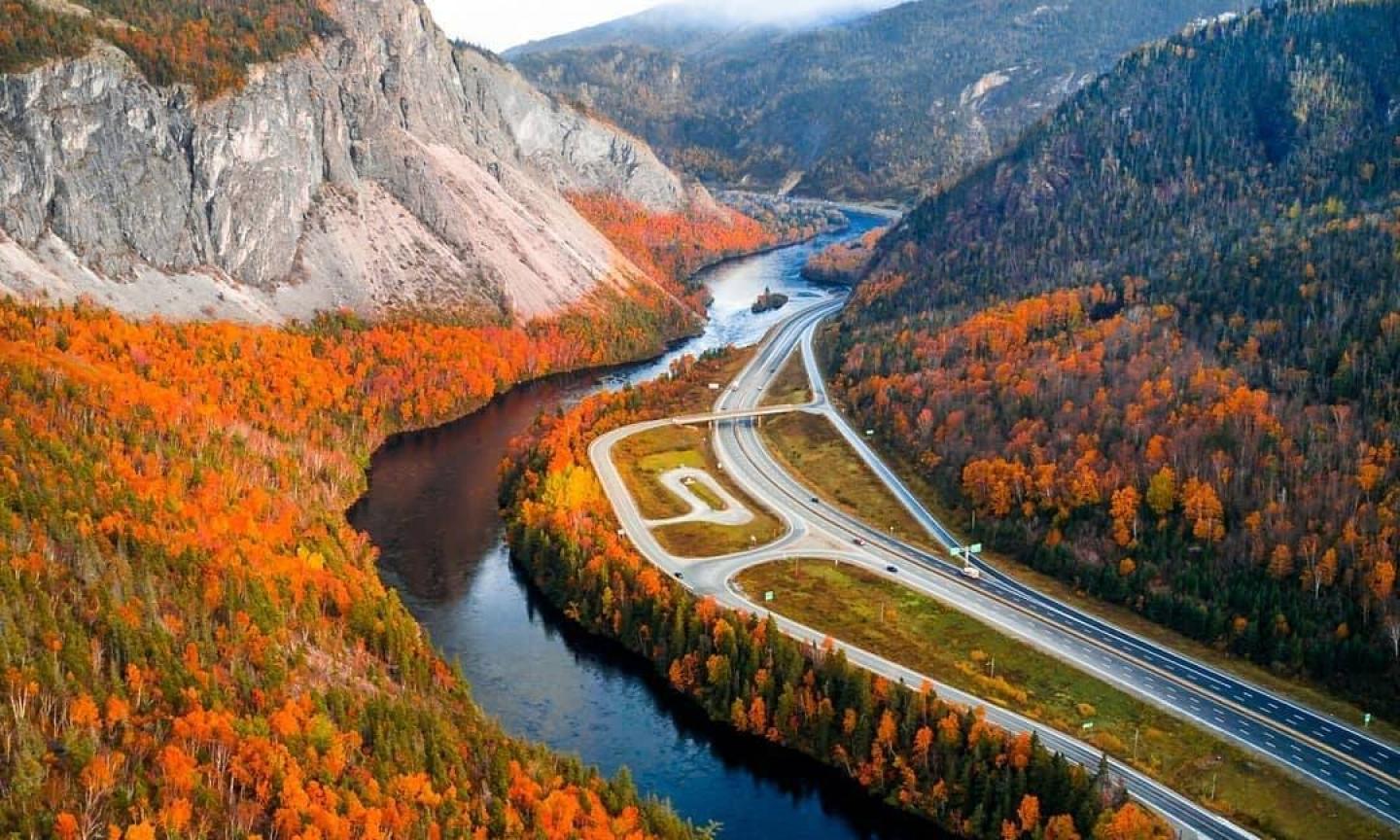 An aerial view of a highway and river winding through a valley blanketed in vivid autumn foliage, framed by rocky cliffs and a misty hilltop.
