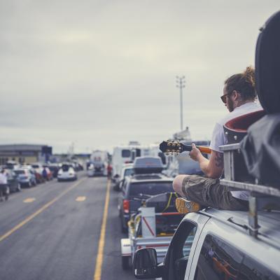 A ponytailed man in his twenties sits on top of a van and plays guitar while waiting to board a Marine Atlantic ferry. 