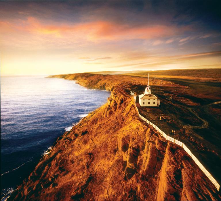 An aerial shot at sunrise shows a white picket fence along a rocky red cliff at the Cape Spear National Historic lighthouse. 