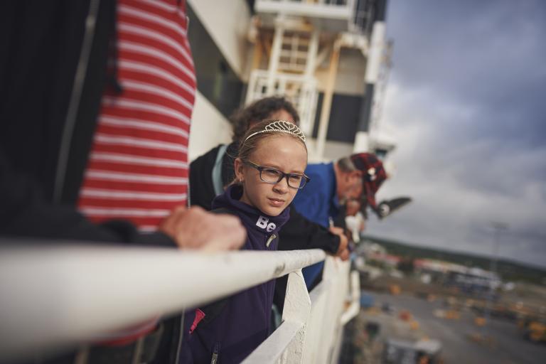 A young girl wearing a tiara and her family look over the white rails of a Marine Atlantic ferry as it leaves port. 