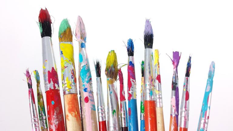 A grouping of art paintbrushes in various sizes, covered in splattered paint colours and set against a white background. 