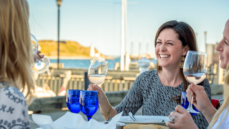 A smiling brunette woman raises a glass of white wine during a patio dinner at a restaurant on the Halifax waterfront. 