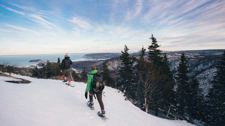 A couple snowshoes past evergreen and birch trees along a snow-covered bluff overlooking mountains, trails, and the Atlantic.