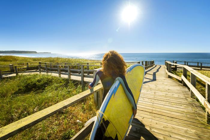 a person with long curly hair in a wetsuit holding a surfboard under their arm stands on wooden boardwalk with ocean in background