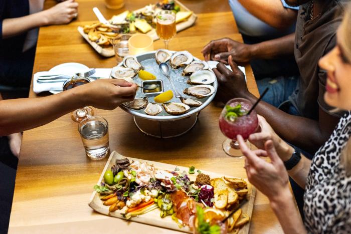 group of people sit around wooden tables with plates of oysters and charcuterie