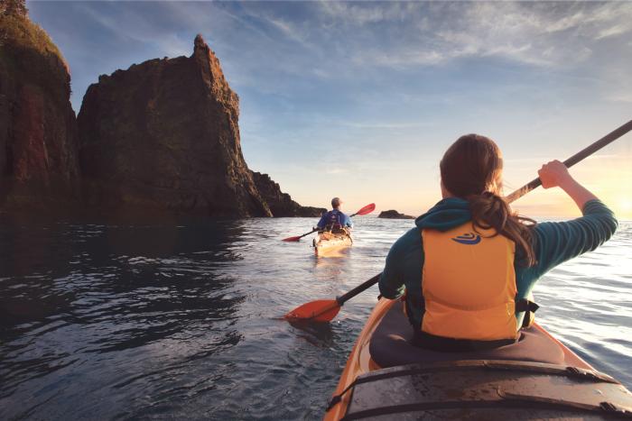 two people in kayaks paddle along still water beside three large rock walls