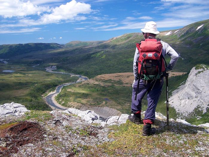 person wearing hat and red backpack stands on edge of lookoff overlooking green wooded area